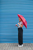 一个年轻的金发女孩拿着一把红伞站在一堵蓝色的墙前。图片素材