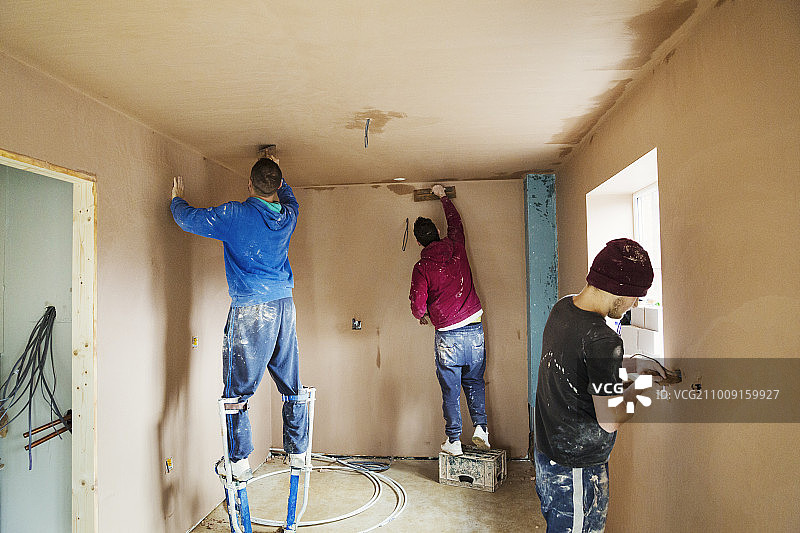 三个人，两个抹灰工踩着高跷，在一所在建的房子的墙上抹平新鲜的灰泥。图片素材
