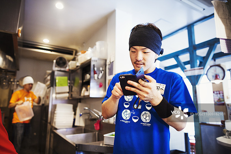 拉面店，员工准备食物。一个厨师在厨房里使用智能手机。图片素材