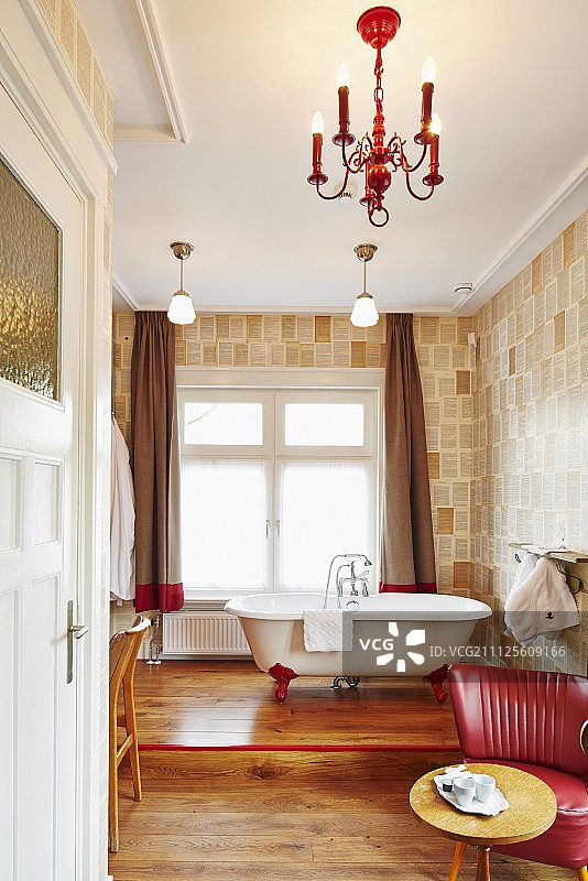 独立式浴缸放在桌子后面的木制平台上，浴室里有红色复古扶手椅和设计师墙纸图片素材