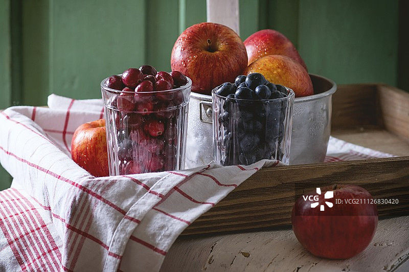 绿色的木墙边，一张旧的白色木椅上放着一碗盛着红苹果和几杯黑莓和越桔的铝碗图片素材