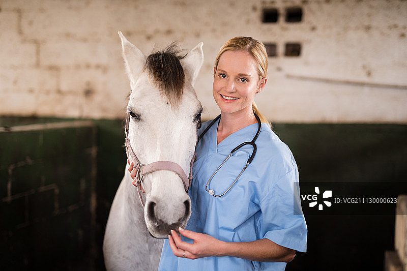 微笑的女兽医站在马在马厩的肖像图片素材