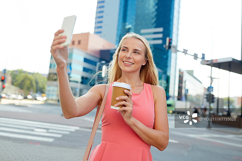 科技、生活方式和人的概念——快乐微笑的年轻女子拿着咖啡和智能手机在城市的街道上自拍。拿着咖啡用智能手机自拍的女人图片素材
