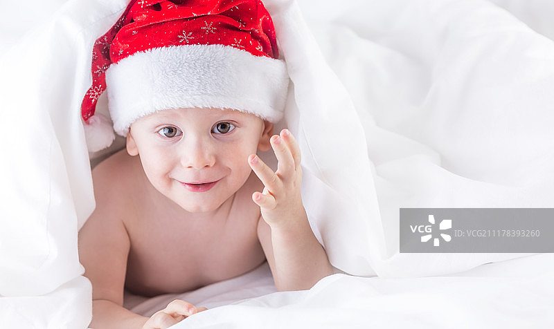 可爱的小男孩戴着圣诞帽，躺在白色羽绒被下的床上图片素材