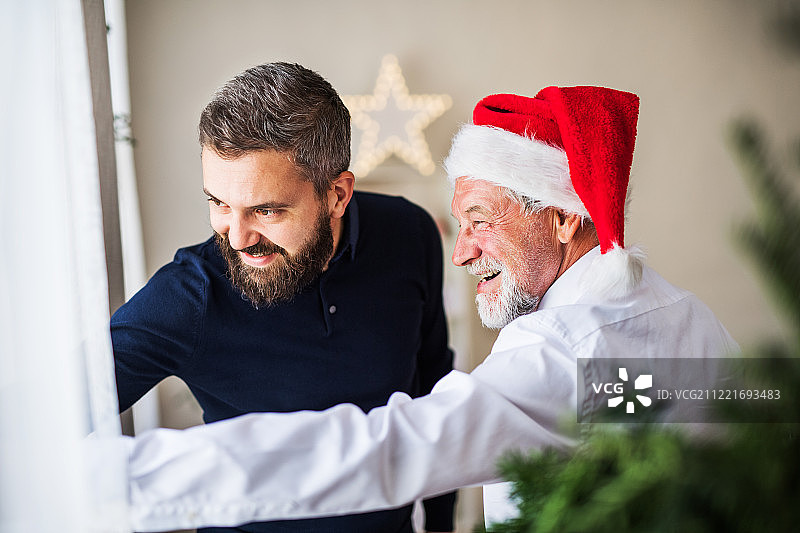 一位戴着圣诞老人帽的年长父亲和已成年的儿子站在家里的窗户旁。图片素材
