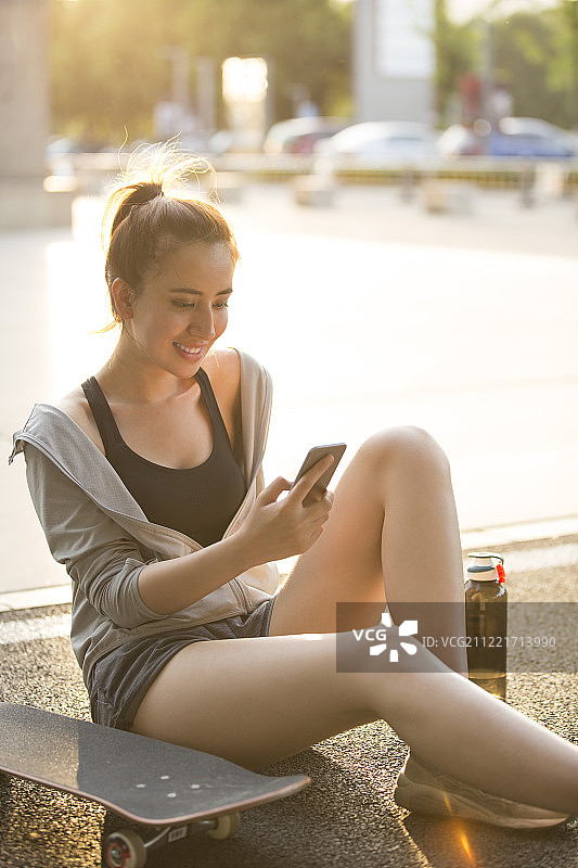 一个年轻女孩在户外滑板运动后坐在地上玩手机休息图片素材
