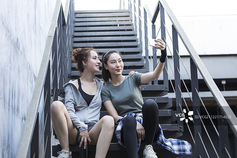 两个青年女子在户外运动后坐在台阶上休息用手机自拍图片素材