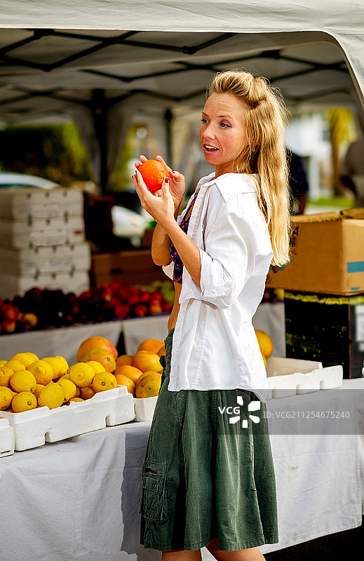 一个金发女人拿着一个橙子站在市场摊位旁，美国格尔夫波特图片素材