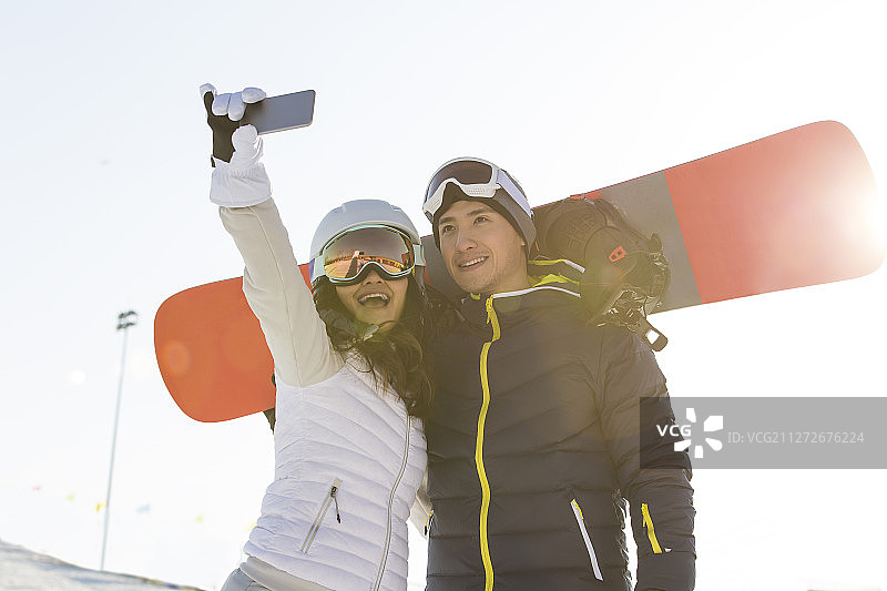 甜蜜情侣背着滑雪板在户外滑雪场拿手机自拍图片素材
