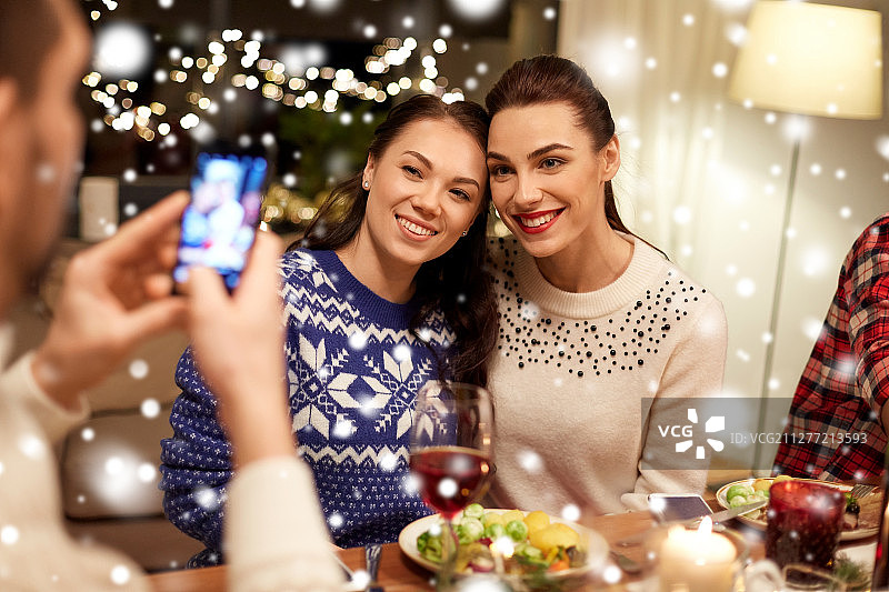 节日和庆祝概念-快乐的朋友在家里吃圣诞晚餐，并通过智能手机在雪中拍照。朋友们在吃圣诞晚餐，拍照图片素材