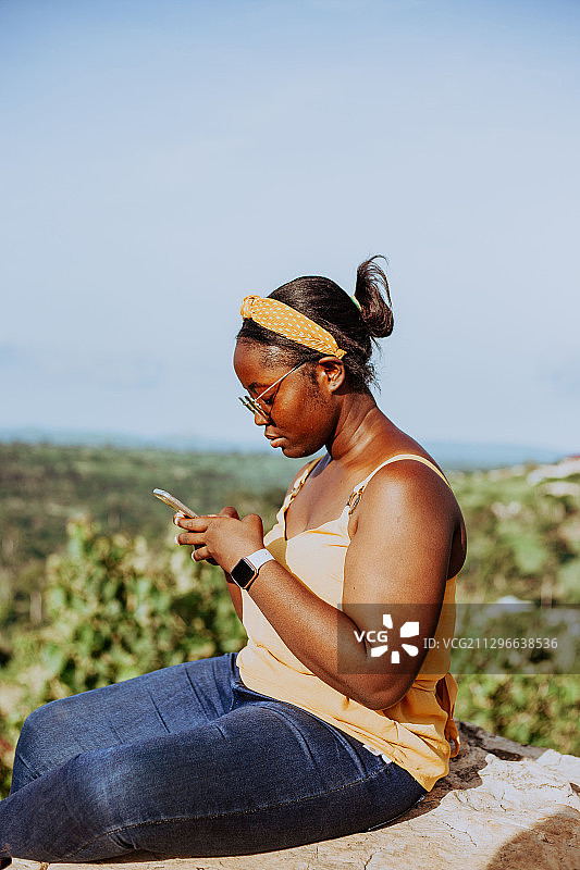 加纳，Mpraeso，一名黑人妇女坐在岩石上玩手机图片素材