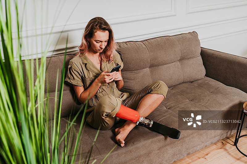 年轻女子戴着假肢坐在家里的沙发上使用手机图片素材