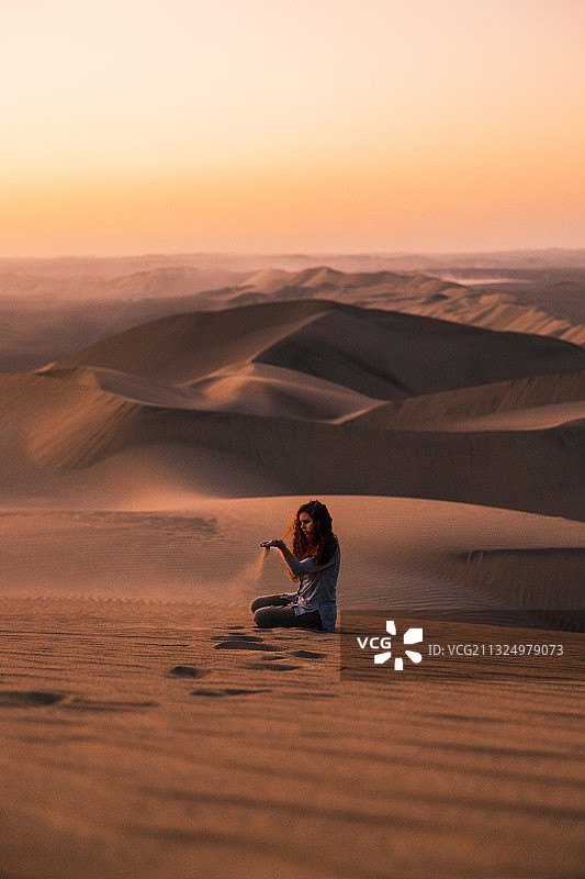 秘鲁伊卡，一名年轻女子坐在日落时的沙漠沙丘上图片素材