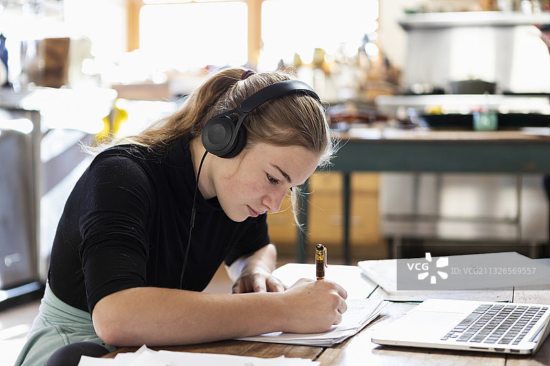 家，美国，十几岁的女孩戴着耳机使用笔记本电脑和在笔记本上写图片素材