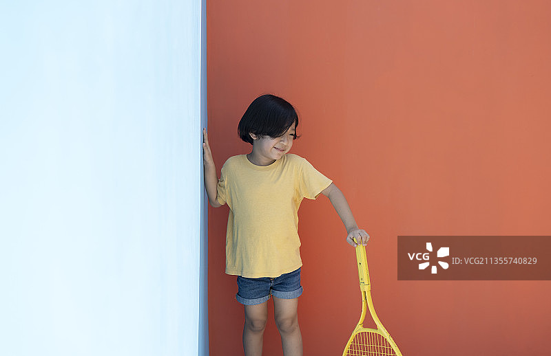 可爱的小男孩穿着黄色的衬衫，手拿网球拍，背景是鲜艳的蓝色和红色。拿着网球拍的害羞男孩。图片素材