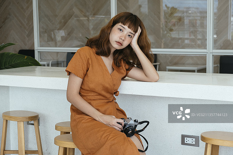 漂亮的年轻亚洲潮女卷发拿着相机坐在椅子上的柜台书桌。图片素材