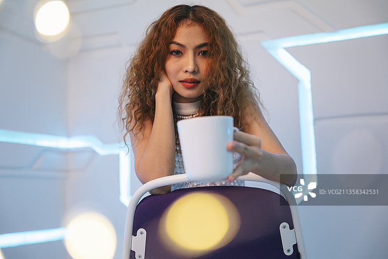 时髦的亚洲卷发女人的肖像坐在椅子上，拿着陶瓷咖啡杯在现代的房间与黄色焦距。图片素材