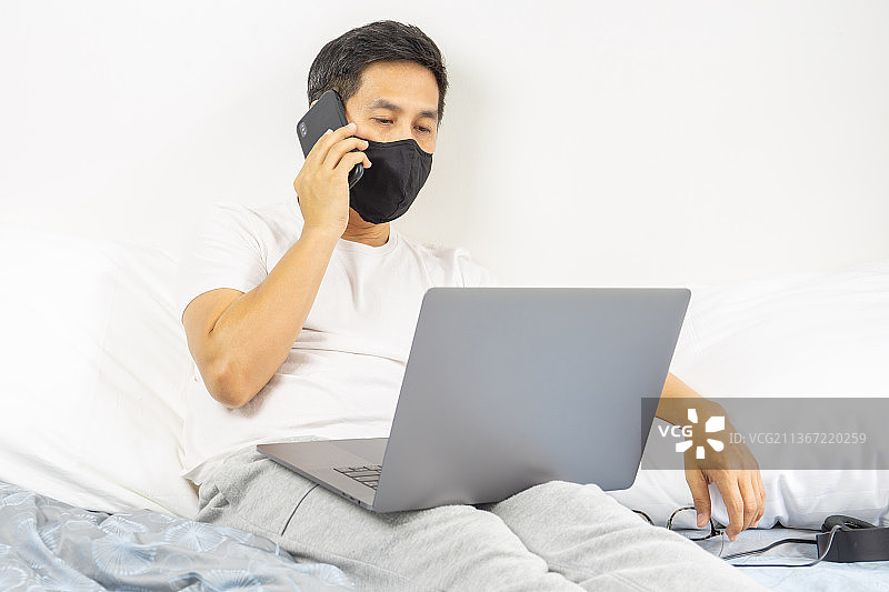 泰国，一名男子戴着防护口罩，膝上放着笔记本电脑打电话图片素材