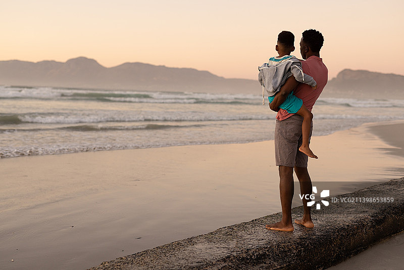一个非洲裔美国年轻人背着儿子站在海滩上的岩石上，夕阳西下，天空晴朗图片素材