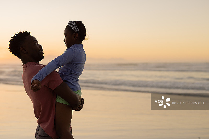 非洲裔美国年轻人在日落时背靠大海和晴朗的天空抱起女儿图片素材