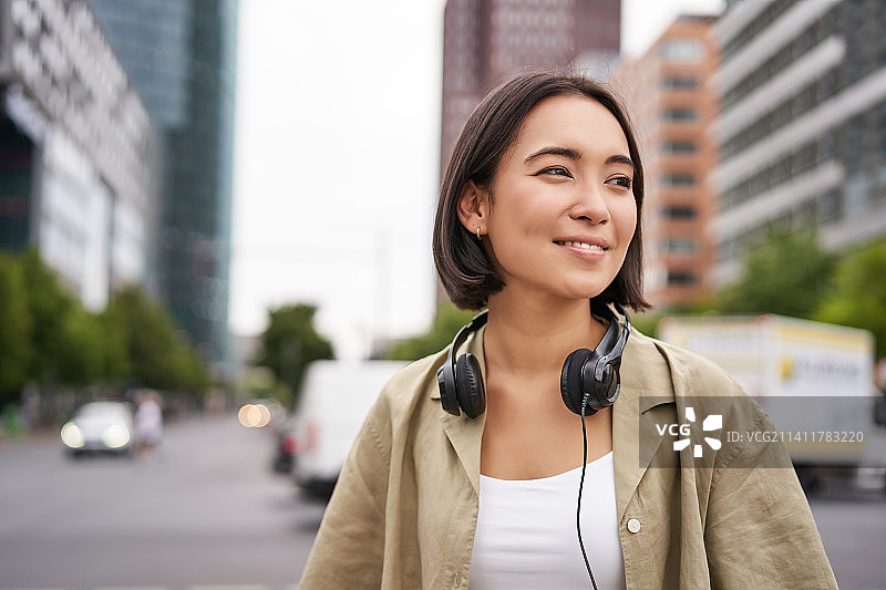 年轻的亚洲女性戴着耳机的肖像，在城市里摆姿势，微笑着图片素材