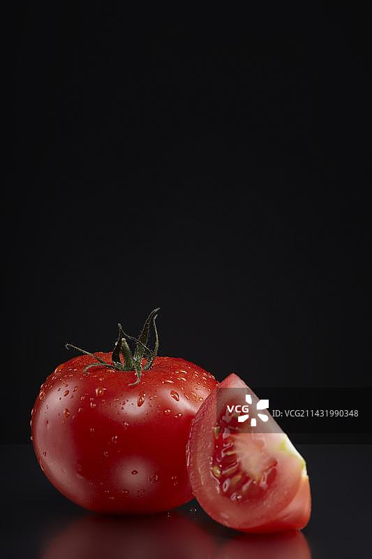 一个新鲜的番茄(Solanum lycopersicum)和一片带水滴的番茄片放在一块深色玻璃盘子上图片素材