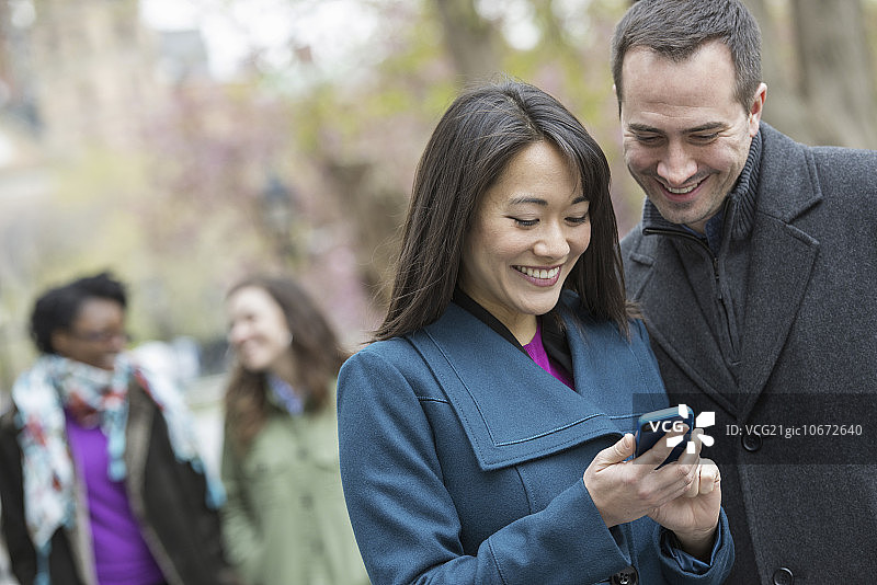 一群人在城市公园里。一个穿灰色外套的男人，和一个穿绿松石外套的女人，都在看智能手机。图片素材