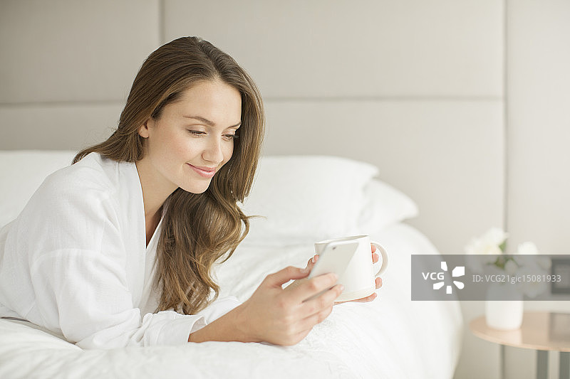 穿着浴袍的微笑女人喝着咖啡，在床上用手机发短信图片素材