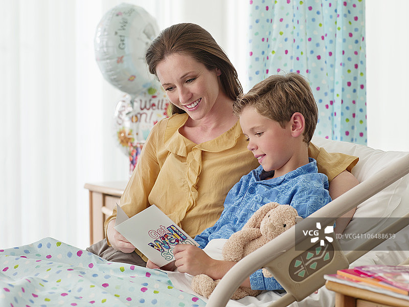 微笑的母亲和儿子在床上(10-11)读“康复”卡在医院图片素材