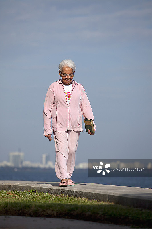 一位年长的女士拿着一本书走在海边的小路上图片素材