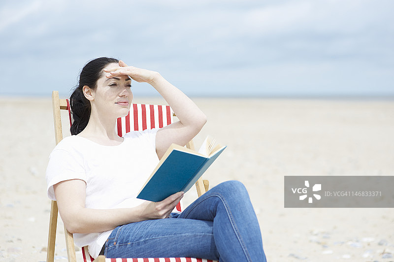 拿着书的女人望着海滩。图片素材