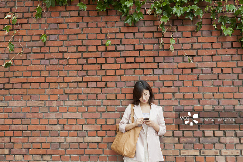 一名年轻女子在用智能手机，倚着砖墙图片素材