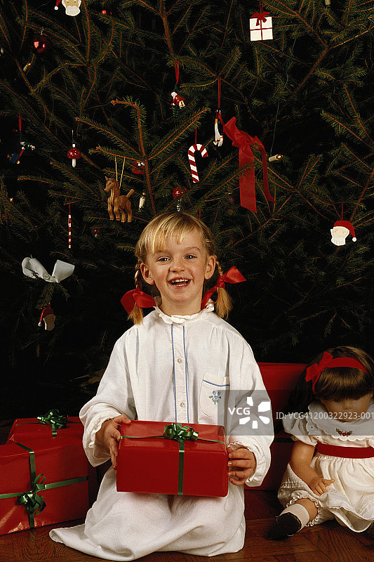 女孩(6-7)带着礼物坐在圣诞树旁，微笑着图片素材