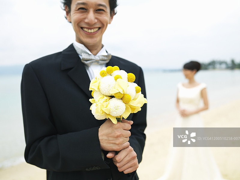 在沙滩上，新郎手持一束鲜花，背景是新娘图片素材