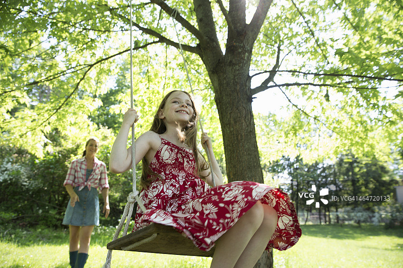 夏天。一个穿着太阳裙的女孩在树枝上荡秋千。她后面有个女人。图片素材