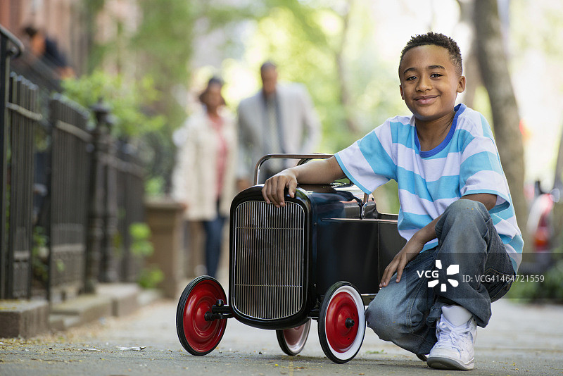 一个小男孩在城市的街道上玩一辆老式的有轮子的玩具车。一对夫妇在一旁观看。图片素材