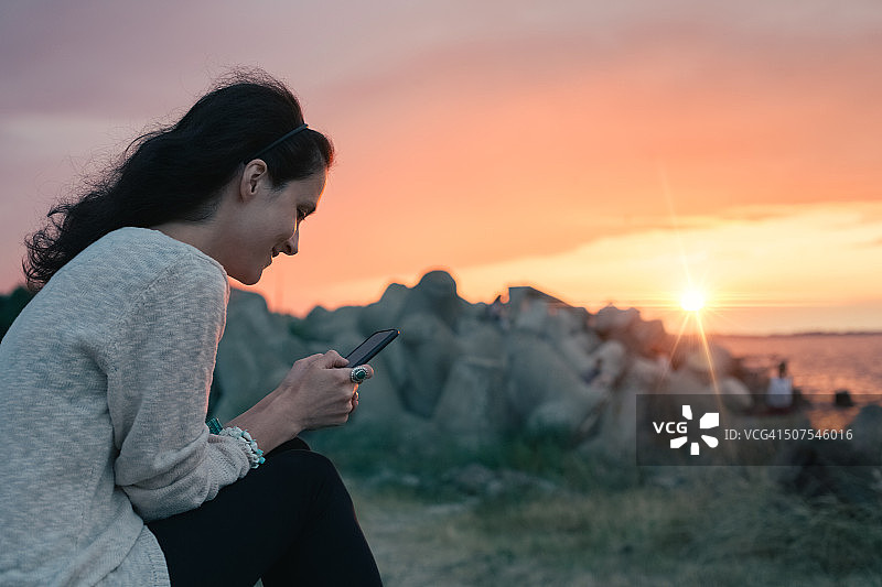 日落时分，岩石海滩上的女孩在用智能手机图片素材