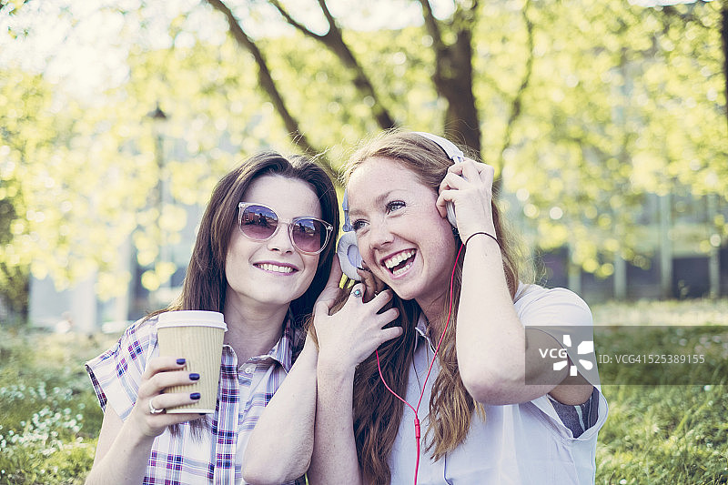 两个年轻的女性朋友在公园里喝咖啡，戴着耳机听音乐图片素材