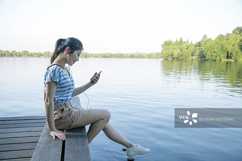 一个年轻的亚洲女人在湖边的码头上看她的手机图片素材