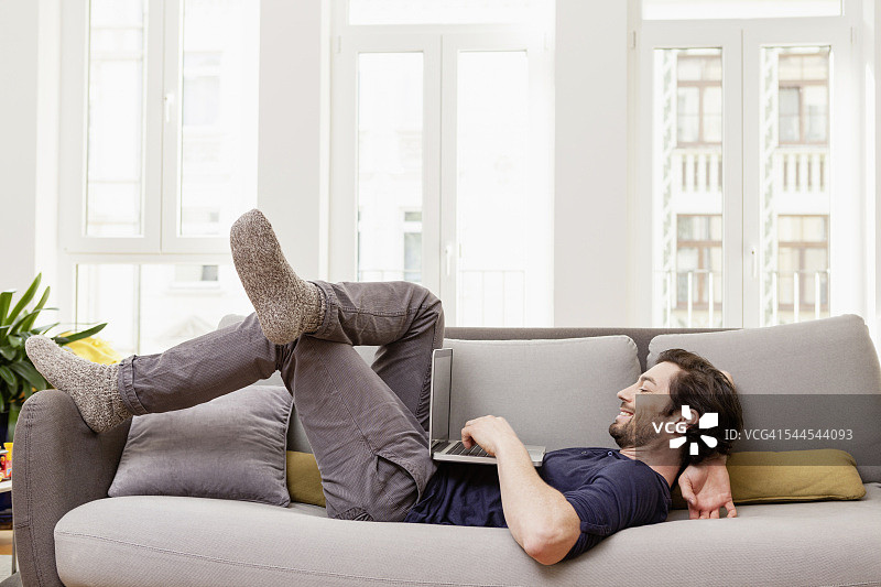 放松的男人躺在沙发上使用笔记本电脑图片素材