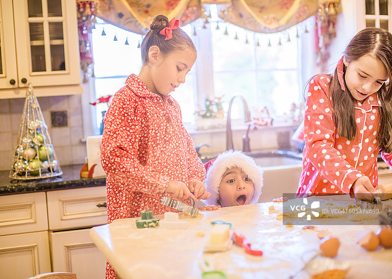 厨房里的男孩戴着圣诞老人帽做饼干，姐妹们从厨房柜台上张大嘴巴偷看图片素材