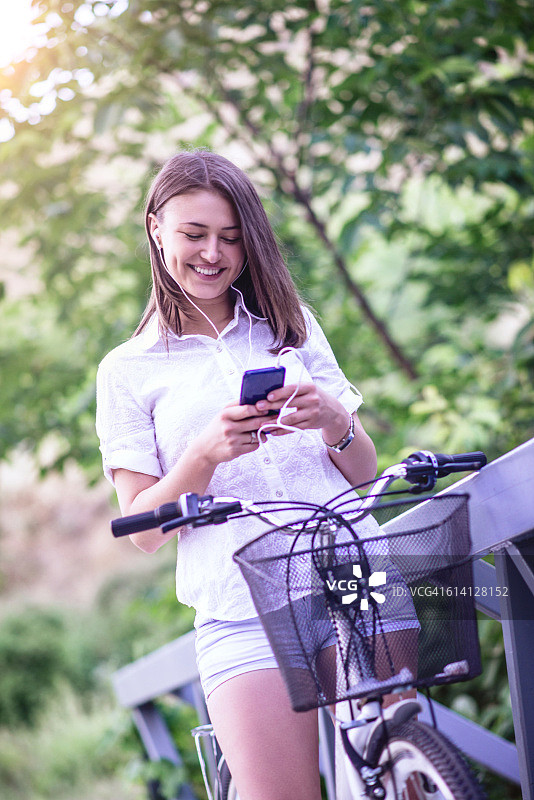 微笑的女人骑自行车和使用智能手机和耳机图片素材