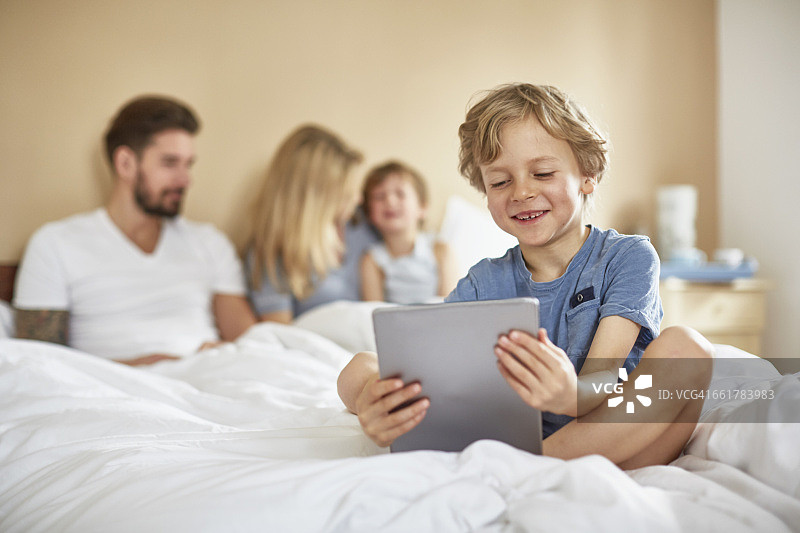 男孩在父母的床上用平板电脑微笑图片素材