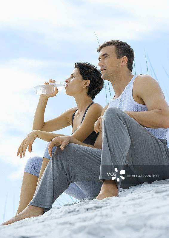 夫妇穿着运动服坐在海滩上，女人喝着水图片素材