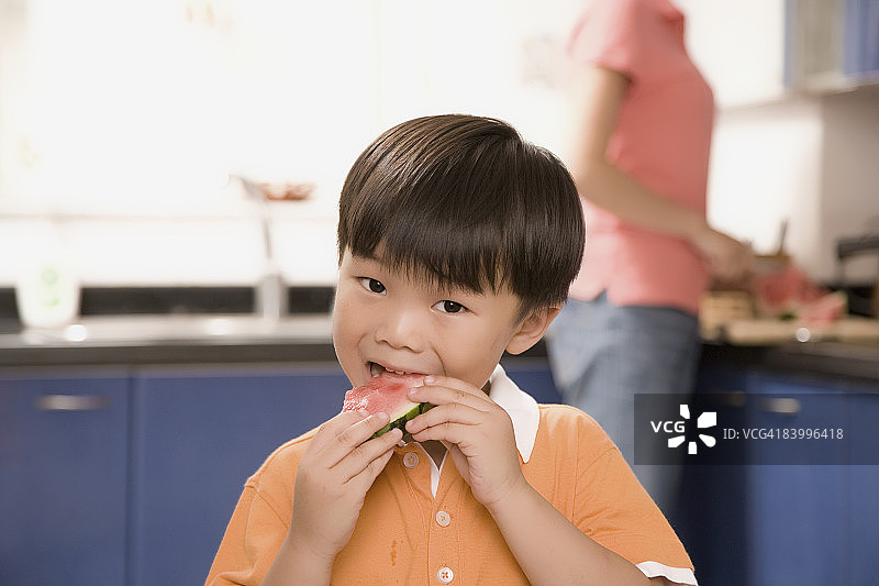 一个男孩吃西瓜的特写镜头，他的妈妈站在他身后图片素材