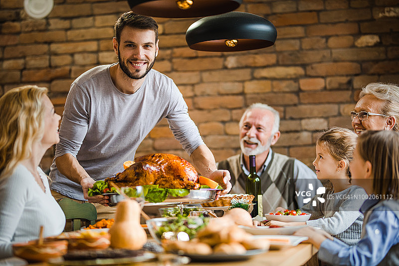 年轻快乐的男人在感恩节餐桌上为他的家人提供填充火鸡。图片素材