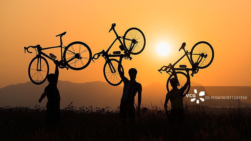 日落时分，三个骑自行车的人把自行车举过头顶的剪影。图片素材