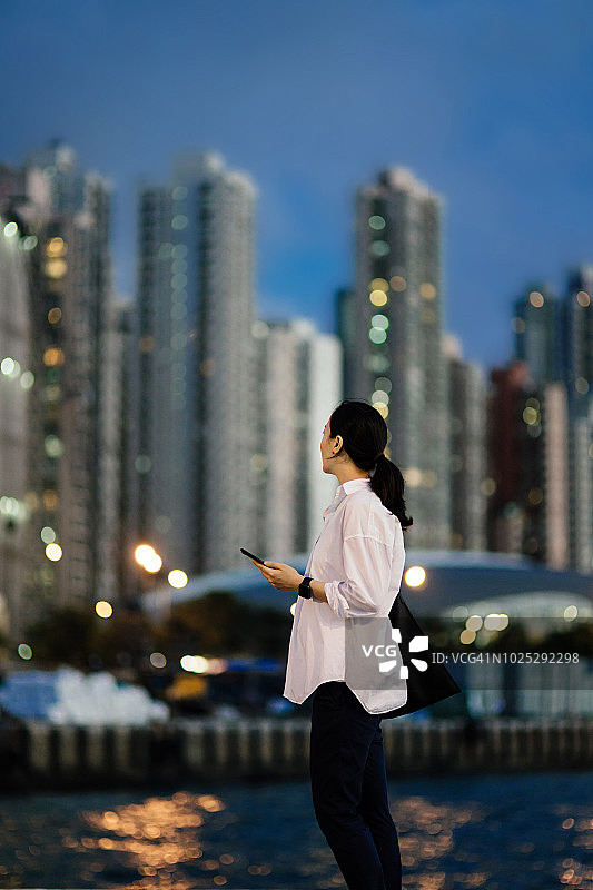 美丽的亚洲女人与智能手机看发光的城市摩天大楼，享受壮观的城市夜景图片素材