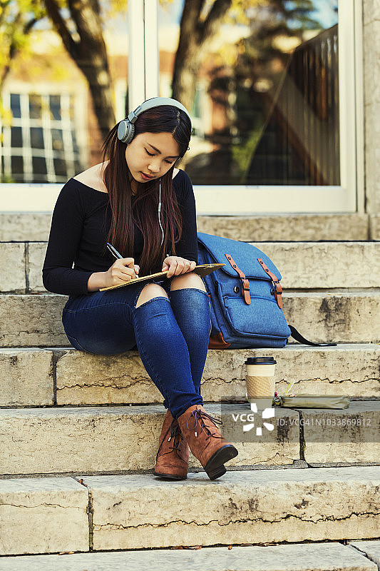 一位年轻的中国女大学生坐在台阶上，戴着耳机听音乐，在笔记本上写字图片素材