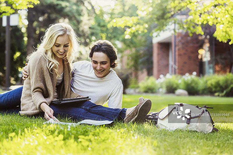 一对年轻夫妇在大学校园的户外草地上学习，一边使用平板电脑一边翻阅课本图片素材